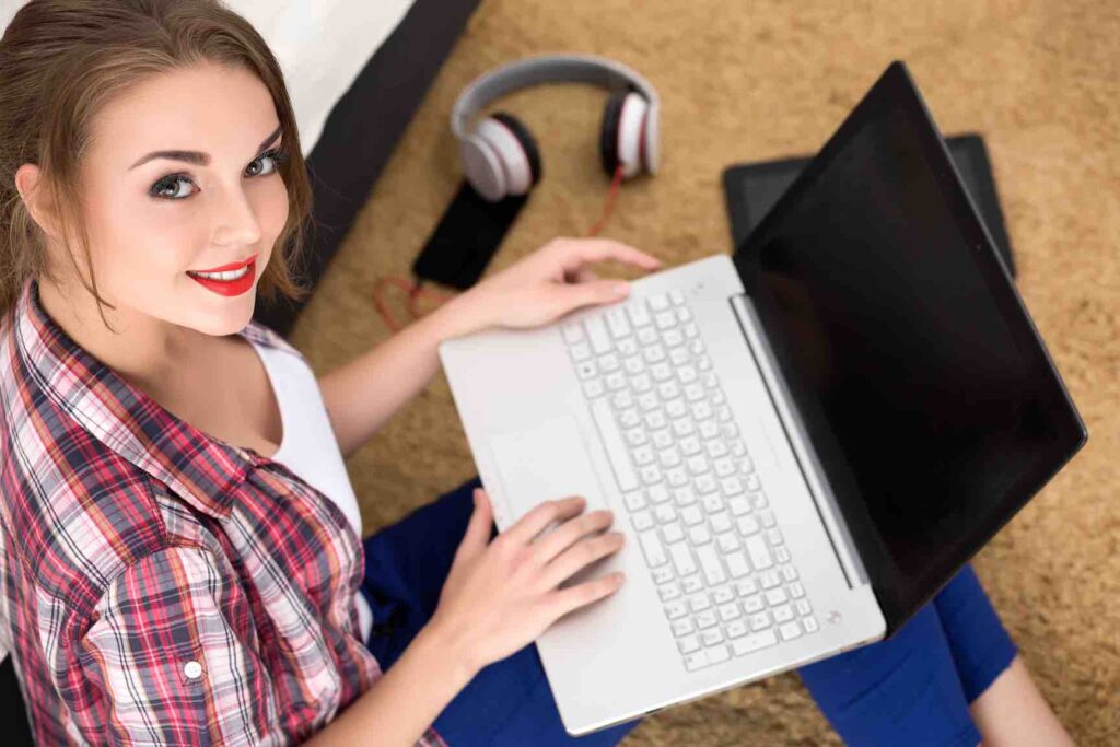 A young woman is smiling, looking up towards the viewer. A laptop is open on her legs and in the background a phone and headphones lie on a brown carpet.