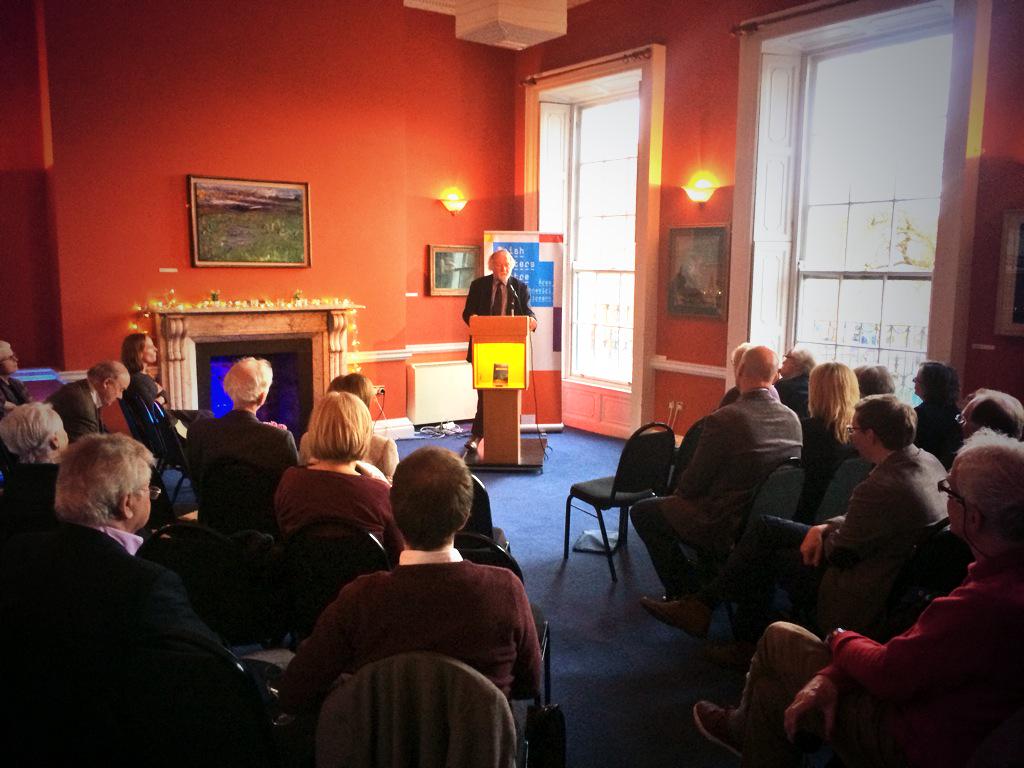 A group of seated people are looking towards literary agent Jonathan Williams who is speaking from a lectern. Jonathan Williams gave advice on what agents are looking for to the Irish Writers Union. The room has paintings hanging on orange walls and feels warm thanks to the glow of two yellow lights.