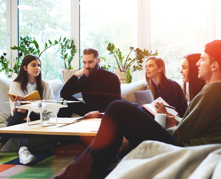 Five young people (F, M, F, F, M) are sitting around a table, four with notebooks and pens, one looking thoughtful. It is a writing workshop. The background is of a modern room with plenty of light from the tall windows behind them.