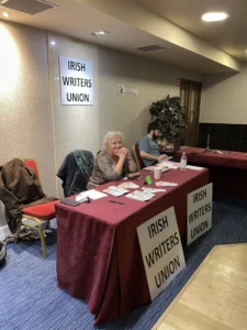Katherine sits alone at a table smiling, with three placards saying Irish Writers Union.